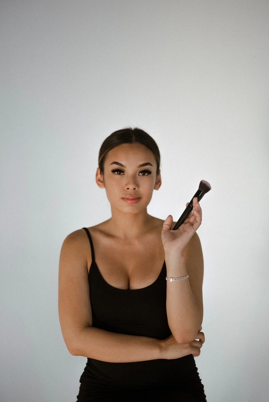 Kira Sarifa, makeup artist, holding a makeup brush in a black tank top against a soft grey backdrop.